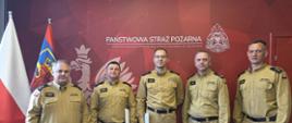 Pięciu strażaków w piaskowych mundurach służbowych stoi w rzędzie na tle czerwonej ściany z logiem Państwowej Straży Pożarnej w Szczecinie. Po lewej stronie widoczne są flagi: Polski oraz flaga z godłem Szczecina. Dwaj strażacy w środku trzymają białe teczki z dokumentami.