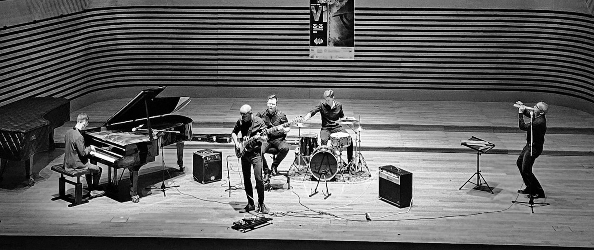 Na estradzie sali koncertowej po lewej stronie na fortepianie gra mężczyzna, na środku stoi mężczyzna grający na gitarze elektrycznej , po prawej przy mikrofonie stoi mężczyzna i gra na dwojnicy. Za nimi siedzi dwóch mężczyzn, jeden gra na gitarze basowej, drugi na zestawie perkusyjnym.