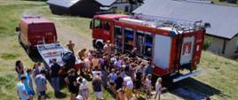 Na zdjęciu strażacy prowadzą pokaz sprzętu ratowniczego dla dzieci i młodzieży w ramach akcji edukacyjnej podczas letniego wypoczynku. Grupa uczestników zgromadziła się wokół wozu strażackiego, z zainteresowaniem oglądając jego wyposażenie. Spotkanie odbywa się w słoneczny dzień, na malowniczym terenie wśród domków i zielonych wzgórz. Na pierwszym planie znajduje się duża grupa dzieci i młodzieży, zgromadzona wokół samochodu ratowniczo-gaśniczego (oznaczenie 511[E]26).