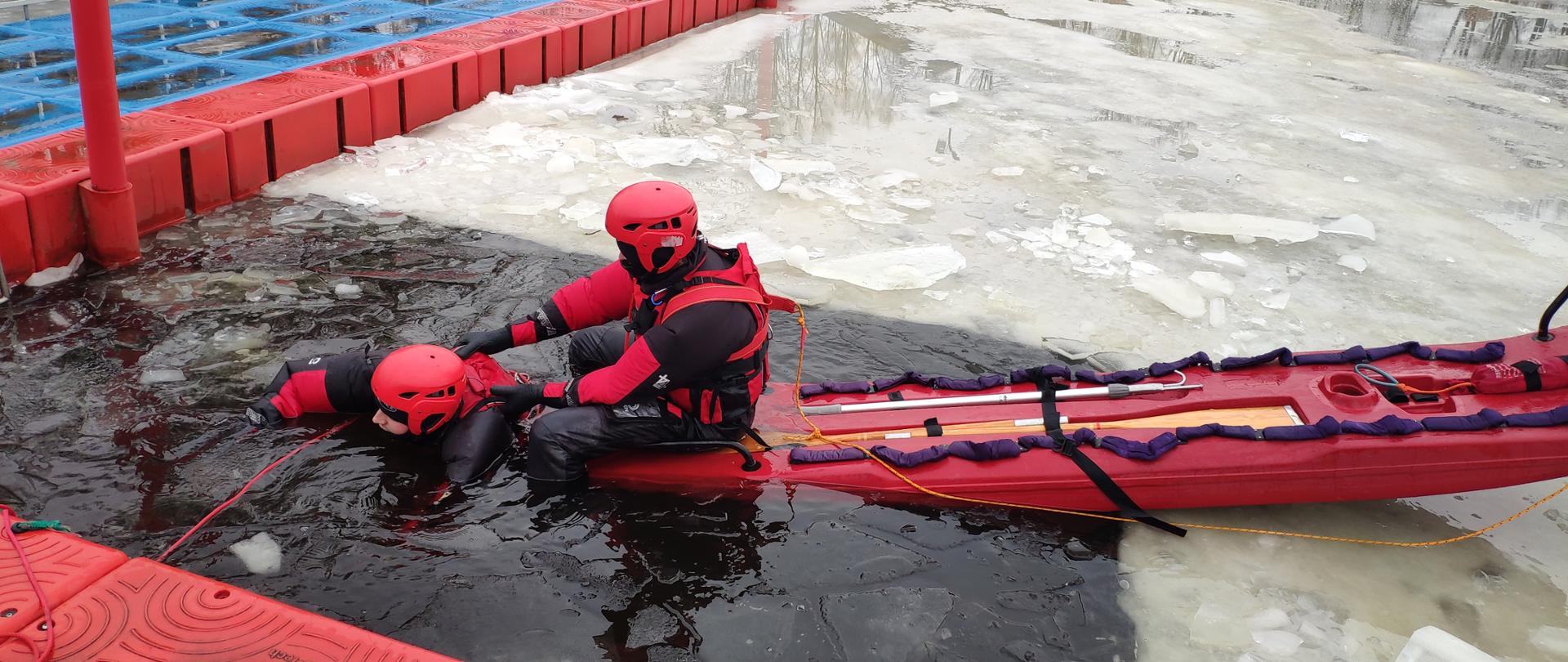 Strażacy ubrani w specjalne kombinezony. Znajdują się w zbiorniku wodnym, dookoła jest zamarznięta woda oraz widać w tle molo. Jeden strażak znajduje się na saniach a drugi w zbiorniku wodnym