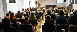 Zdjęcie przedstawia licznie zgromadzonych uczniów w auli szkoły, w tle mgr Agata Bartoś, która prowadzi wykład. Kolorystyka zdjęcia jest brązowo biała. 