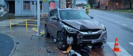 Na zdjęciu widać rozbity ciemny samochód osobowy stojący częściowo na jezdni a częściowo na chodniku. Pojazd, który uderzył w barierki, znak i słup sygnalizacji świetlnej ma rozbity przód. W tle widać strażaka w żółtym hełmie i piaskowym stroju, który kieruje ruchem na drodze, samochody, kamienice, znaki drogowe, latarnie oraz drzewa. Jest widno, ale pochmurnie.