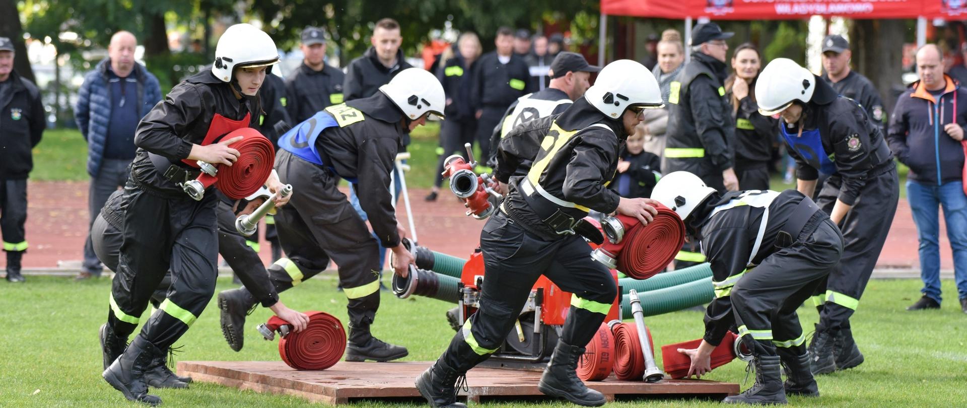Na zdjęciu widać strażaków ubranych umundurowanie koszarowe z emblematami na klatce. Strażacy wykonują ćwiczenie bojowe jedną z konkurencji w zawodach. 