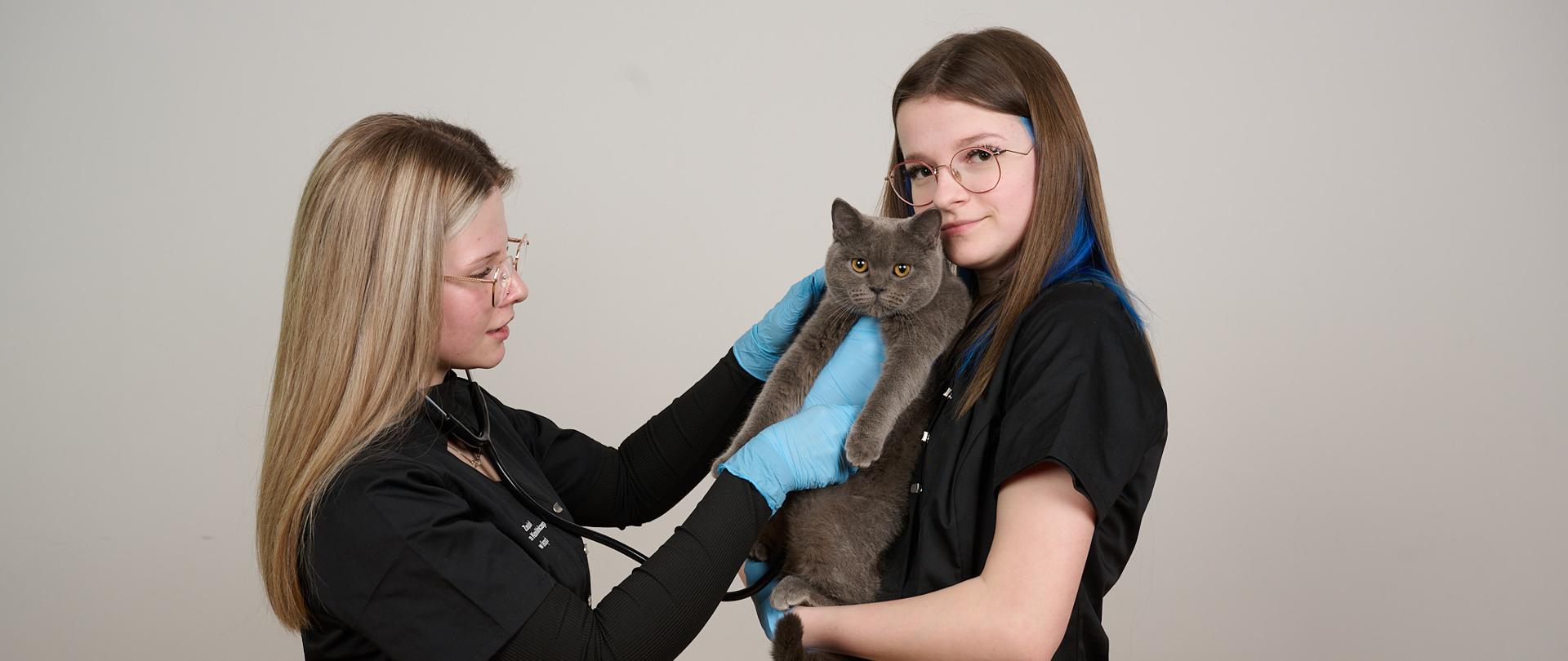 Dwie uczennice stoją zwrócone do siebie. Uczennica z lewej strony stetoskopem bada kota. Druga uczennica trzyma kota na rękach 