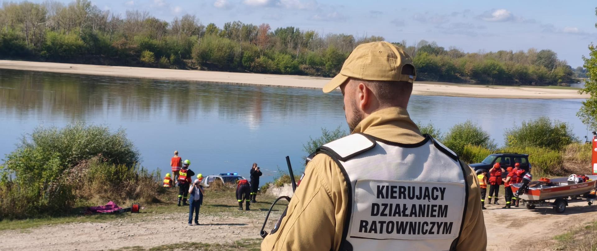 Na pierwszym planie stoi odwrócony plecami strażak w piaskowym mundurze służbowym oraz niebieskiej kamizelce z napisem "KIERUJĄCY DZIAŁANIEM RATOWNICZYM". W tle, na brzegu rzeki widać kilkoro ludzi ubranych w stroje do ratownictwa wodnego, łódkę PSP oraz dwa samochody. Za rzeką widoczna jest plaża i las, a ponad nimi pogodne błękitne niebo.