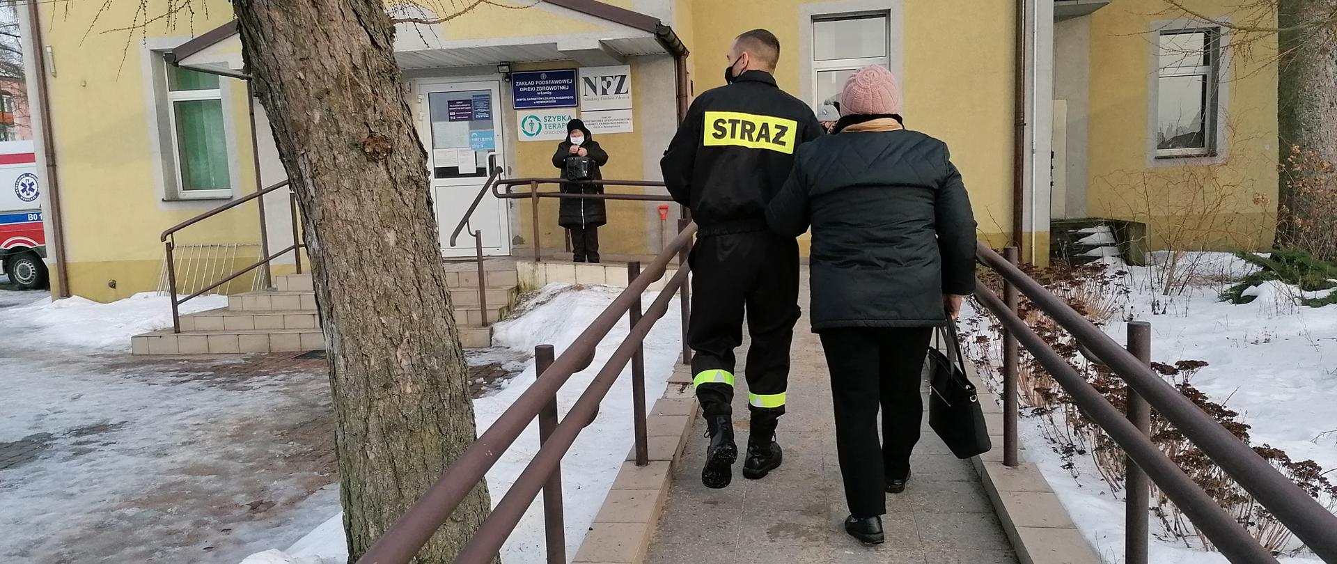 Strażak prowadzi pod rękę osobę starszą do punktu szczepień idąc podjazdem dla osób niepełnosprawnych. W tle widoczny jest punkt szczepień. 