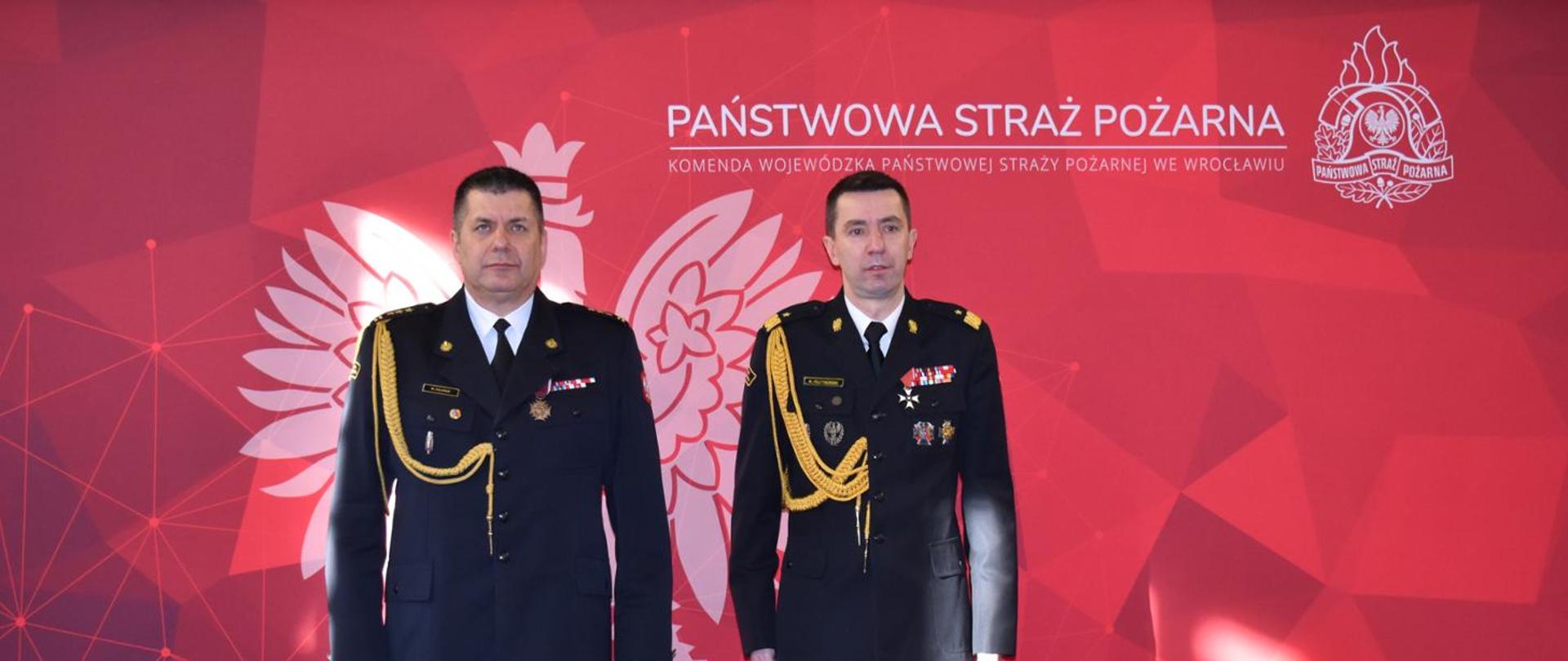 Na tle baneru w Państwowej Straży Pożarnej do zdjęcia pozuje dwóch funkcjonariuszy PSP ubranych w mundury wyjściowe ze sznurem galowym