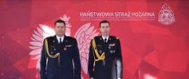 Na tle baneru w Państwowej Straży Pożarnej do zdjęcia pozuje dwóch funkcjonariuszy PSP ubranych w mundury wyjściowe ze sznurem galowym