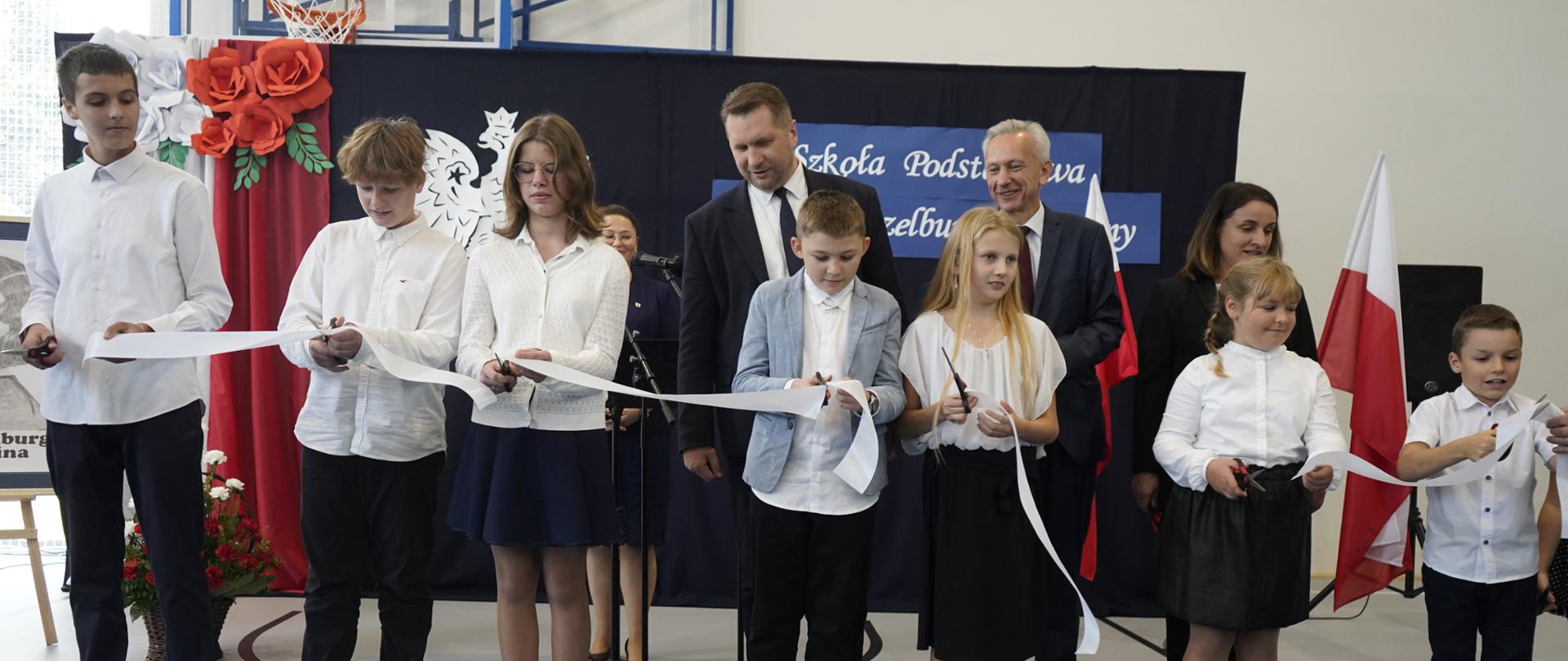 Pod ściana stoi w rzędzie siedmioro dzieci, trzymają nożyczki i przecinają białą rozciągniętą w poprzek przed nimi wstążkę, za nimi stoi minister Czarnek i mężczyzna w garniturze.