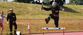 Strażak ubrany w czarne ubranie koszarowe na głowie ma srebrny hełm przeskakuje przez płotek. W tle bieżnia oraz nasyp ziemny porośnięty trawą oraz widzowie