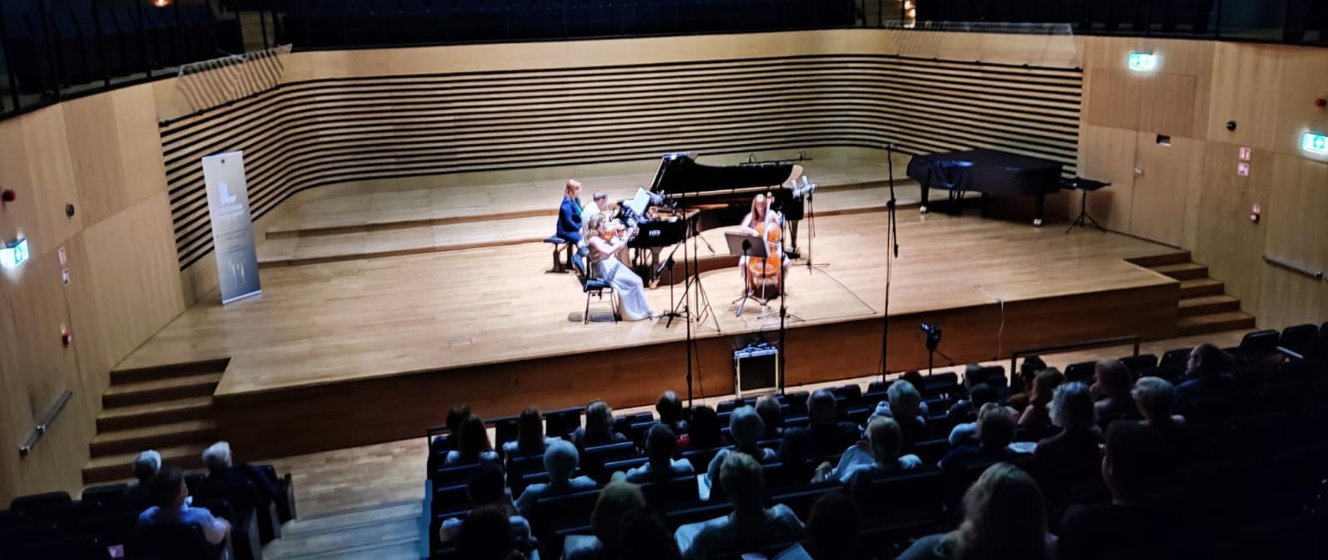 Dwie kobiety siedzą na krzesłach na estradzie i grają na skrzypcach i wiolonczeli, za nimi mężczyzna gra na fortepianie, koło mężczyzny siedzi kobieta. Z przodu widoczna od tyłu publiczność na widowni.