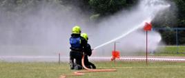 zień. Murawa boiska. Dwóch strażaków ubranych w czarne ubrania i żółte kaski klęczy na murawie. Jeden trzyma proądownicę wodną z której strumieniem wody strąca stojące na metalowych prętach stożki ostrzegawcze.