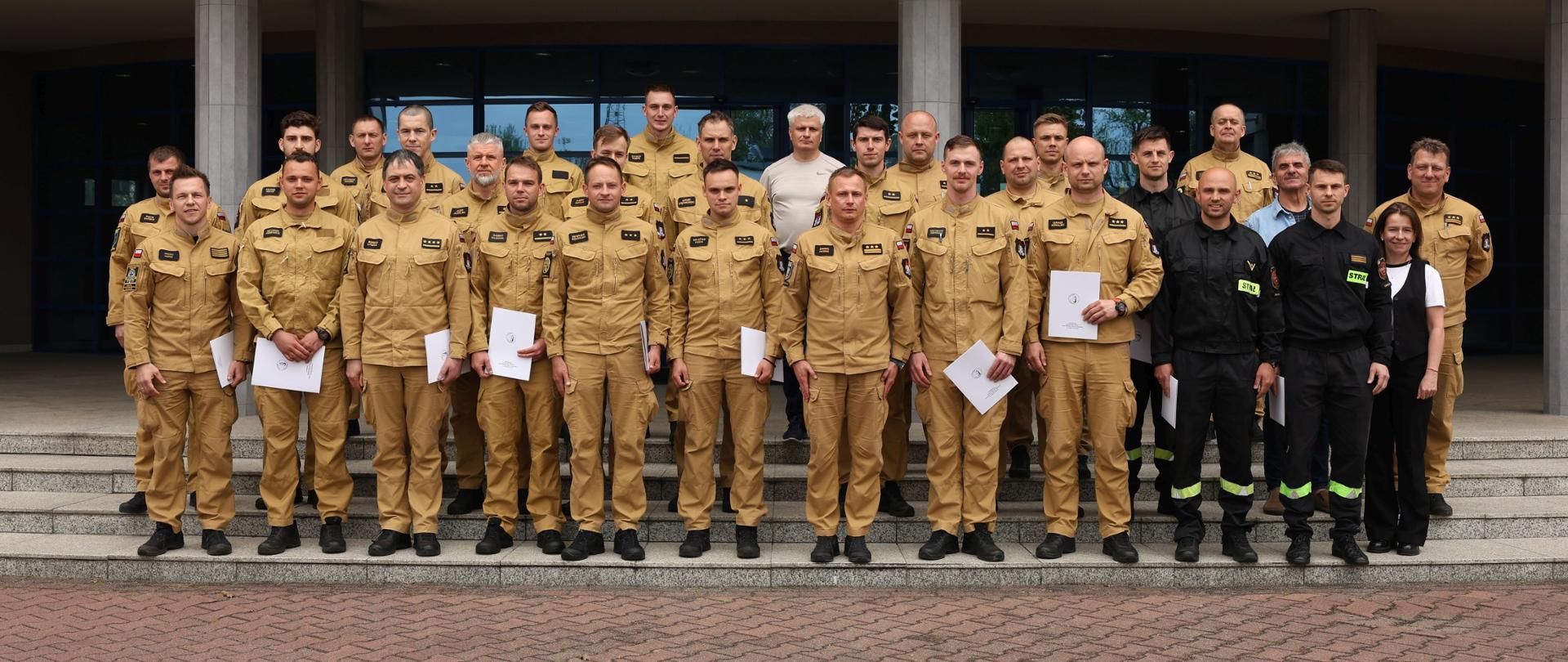 Pamiątkowe zdjęcie grupowe funkcjonariuszy PSP będących uczestnikami szkolenia z zakresu awaryjnego rozładunku cystern (23 osoby) w towarzystwie Zastępcy Komendanta Centralnej Szkoły PSP, kobiety, trzech funkcjonariuszy CS PSP oraz dwóch instruktorów podczas zajęć