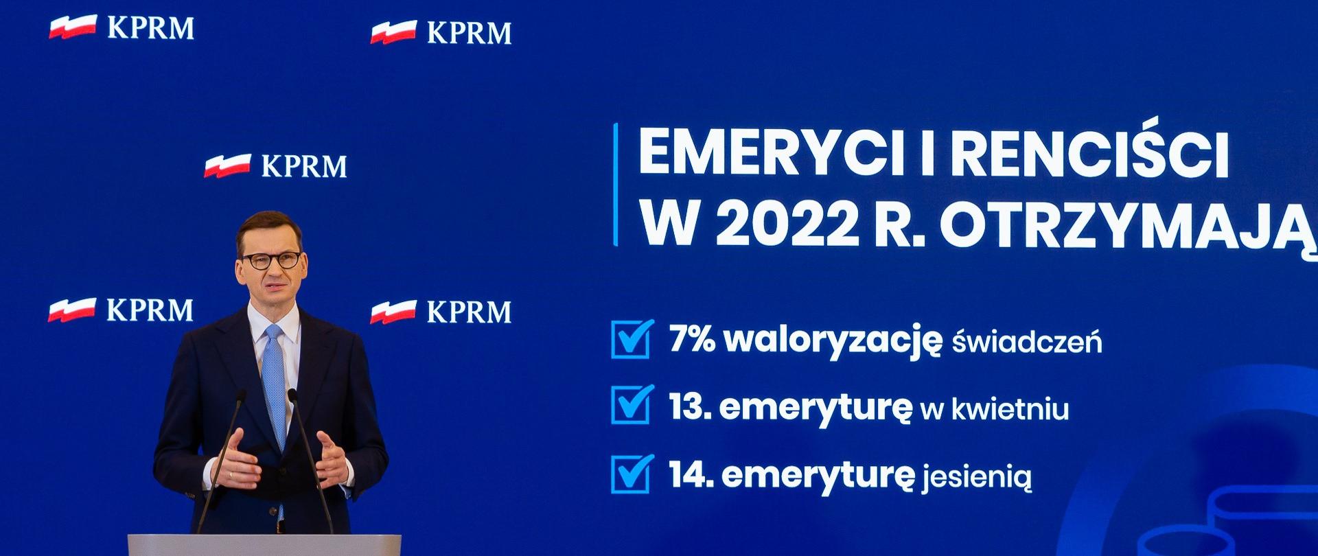 Premier Mateusz Morawiecki podczas konferencji prasowej w KPRM stoi na podeście, przed mównicą, w tle ekran wyświetlający prezentacje