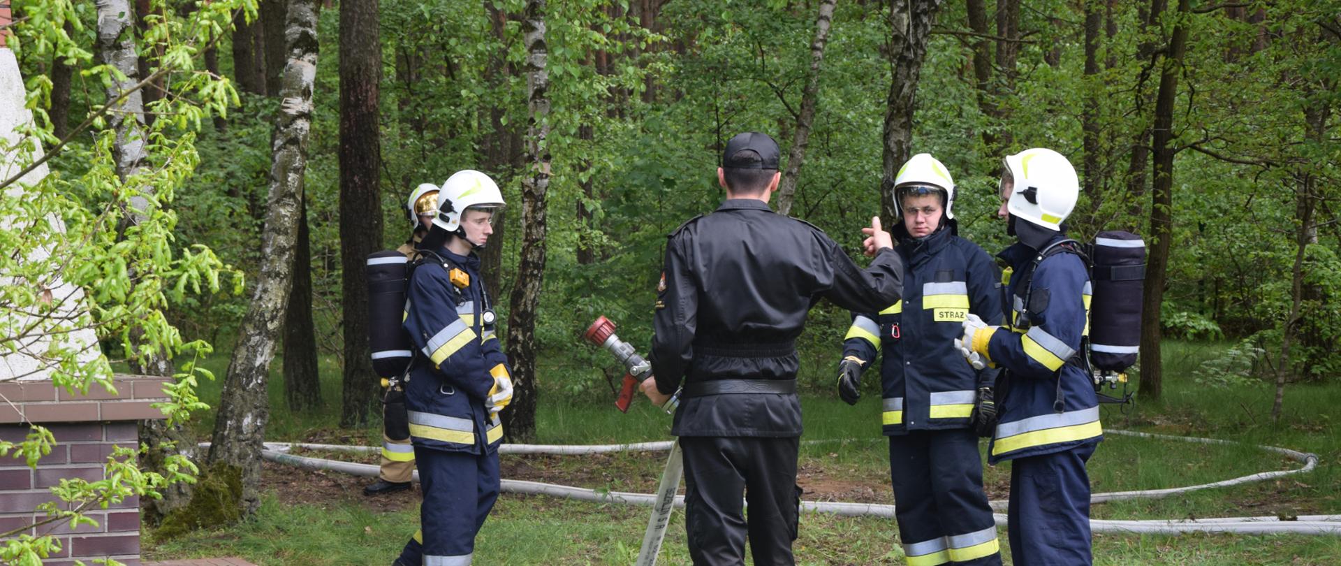 Na zdjęciu widać czterech członków OSP ubranych w ubranie specjalne wraz z egzaminatorem ubranym w mundur. Strażak odwrócony tyłem trzyma w dłoniach wąż z prądownicą i daje wskazówki. Druhowie uważnie słuchają.