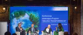 Kadr przedstawia czworo prelegentów podczas konferencji, dwóch mężczyzn i dwie kobiety. Siedzą na fotelach na scenie, a w tle widać duży ekran z napisem "Konferencja otwierająca Program Zielona Transformacja".