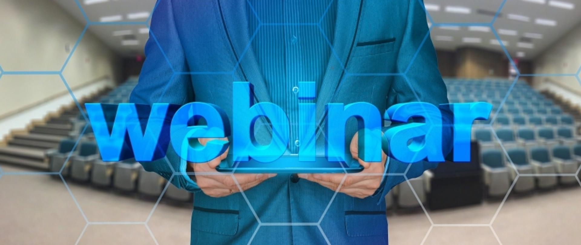 Tablet z napisem „webinar” trzymany przez osobę w garniturze; w tle pusta sala wykładowa i wzory heksagonów.
