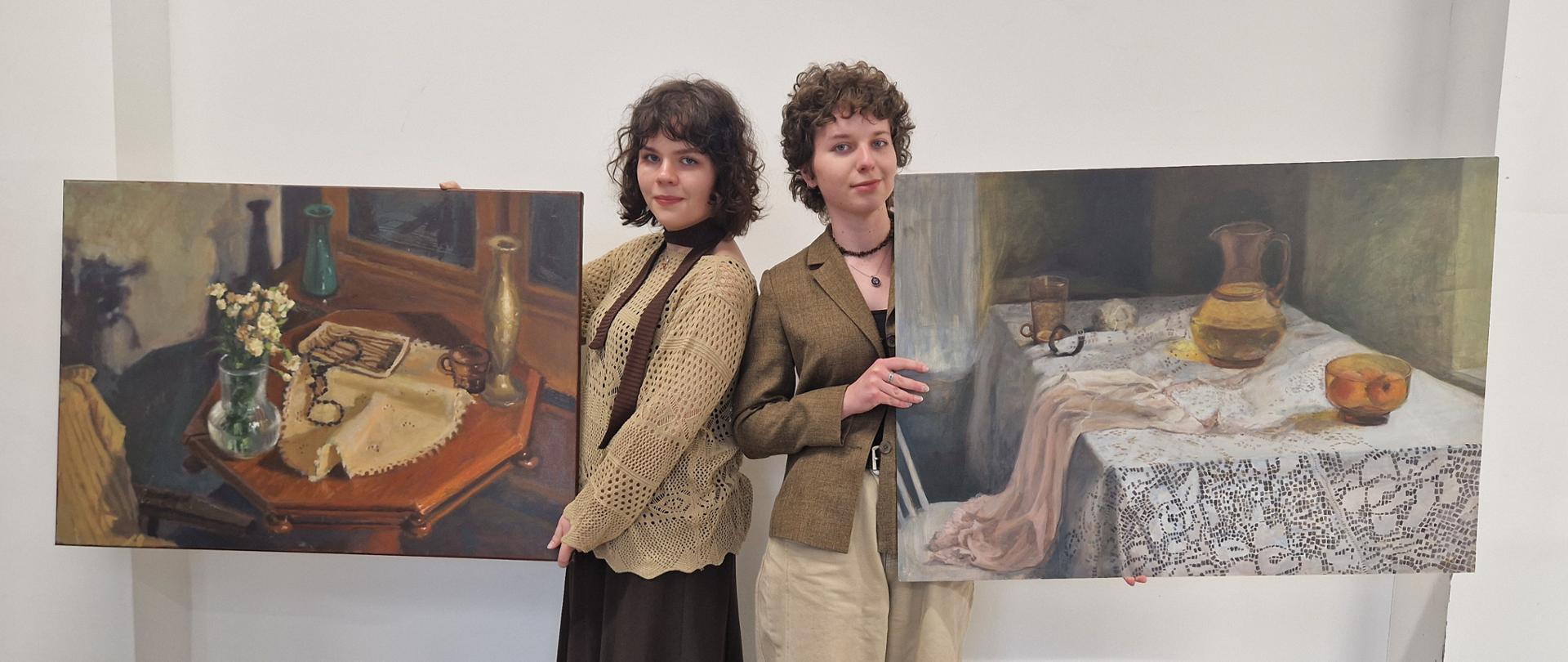 Na zdjęciu widzimy dwie uczennice: Izabelę Sarecką z kl. 4A i Alicję Krzyżańską z kl. 4C. Dziewczyny zajęły 8 i 14 miejsce w konkursie makroregionalnym w Kielcach z rysunku, malarstwa i rzeźby. Konkurs organizowany jest przez Centrum Edukacji Artystycznej w Warszawie i jest konkursem ogólnopolskim. Dziewczyny stoją obok siebie, odwrócone do siebie plecami i w rękach trzymają swoje obrazy. Dziewczyna po lewej stronie Izabela Sarecka ma krótkie, czarne i kręcone włosy, spogląda na nas i się uśmiecha. Jest ubrana w beżowy sweter i brązową spódnicę i brązowe buty. Dziewczyna po prawej stronie ma również ciemne, krótkie i kręcone włosy, uśmiecha się do nas. Jest ubrana w brązową marynarkę i beżowe spodnie oraz zielone buty. 