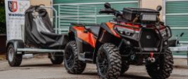 Widok z boku. Przekazywany quad z przypiętą przyczepą do transportu osoby poszkodowanej. W tle boksy garażowe jednostki ratowniczo-gaśniczej. 