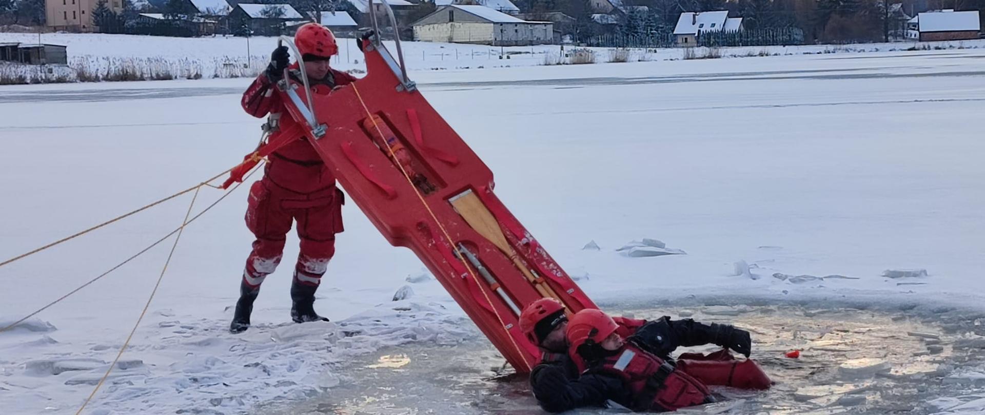 Zdjęcie przedstawia pozorującego topienie się w przerębli odpowiednio zabezpieczonego ratownika, którego ratuje dwóch strażaków. Jeden znajduje się w wodzie asekurując poszkodowanego, a drugi, przy użyciu sań lodowych pomaga ewakuować zagrożoną osobę z wody.