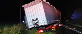 Zdjęcie wykonano porą nocną. Samochód ciężarowy z naczepą w przydrożnym rowie. W tle policjant kierujący ruchem.