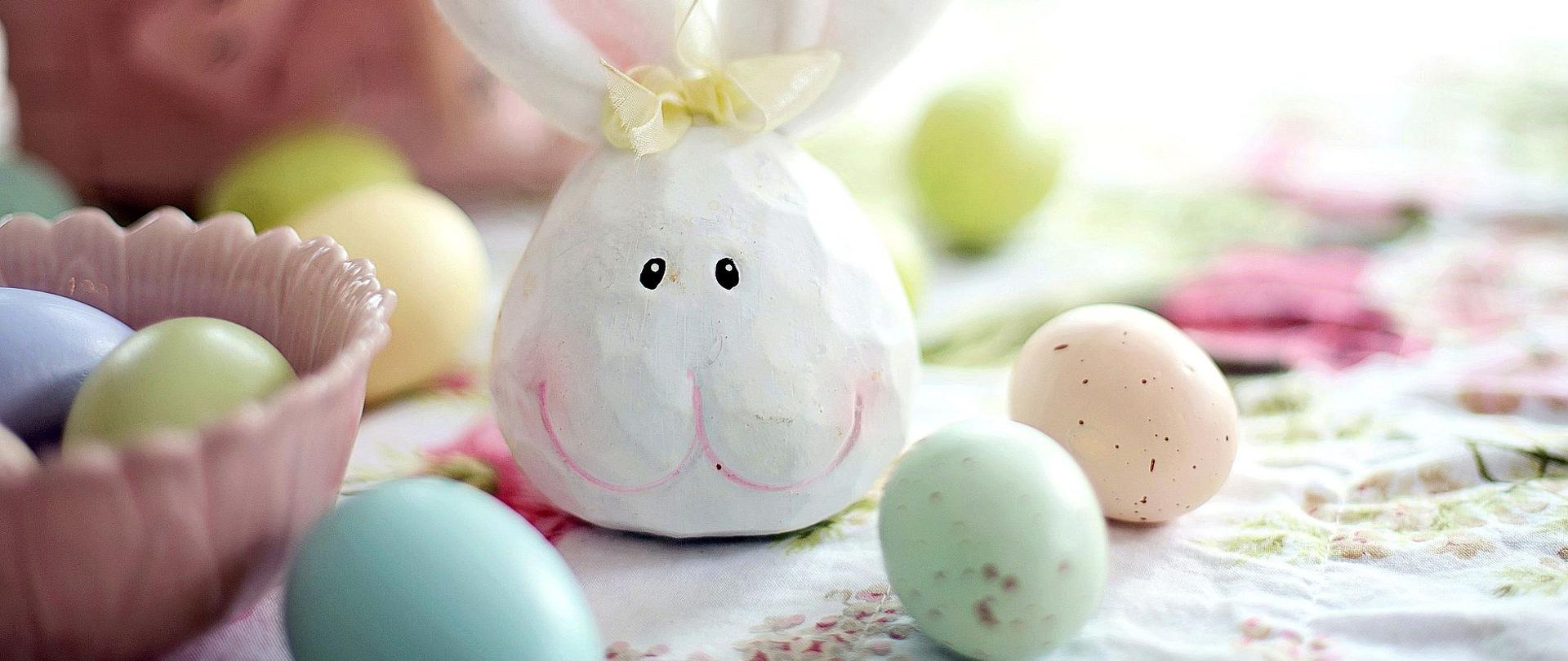 Zdjęcie przedstawia figurkę wielkanocnego zająca oraz pisanek leżących na stole na białym obrusie.