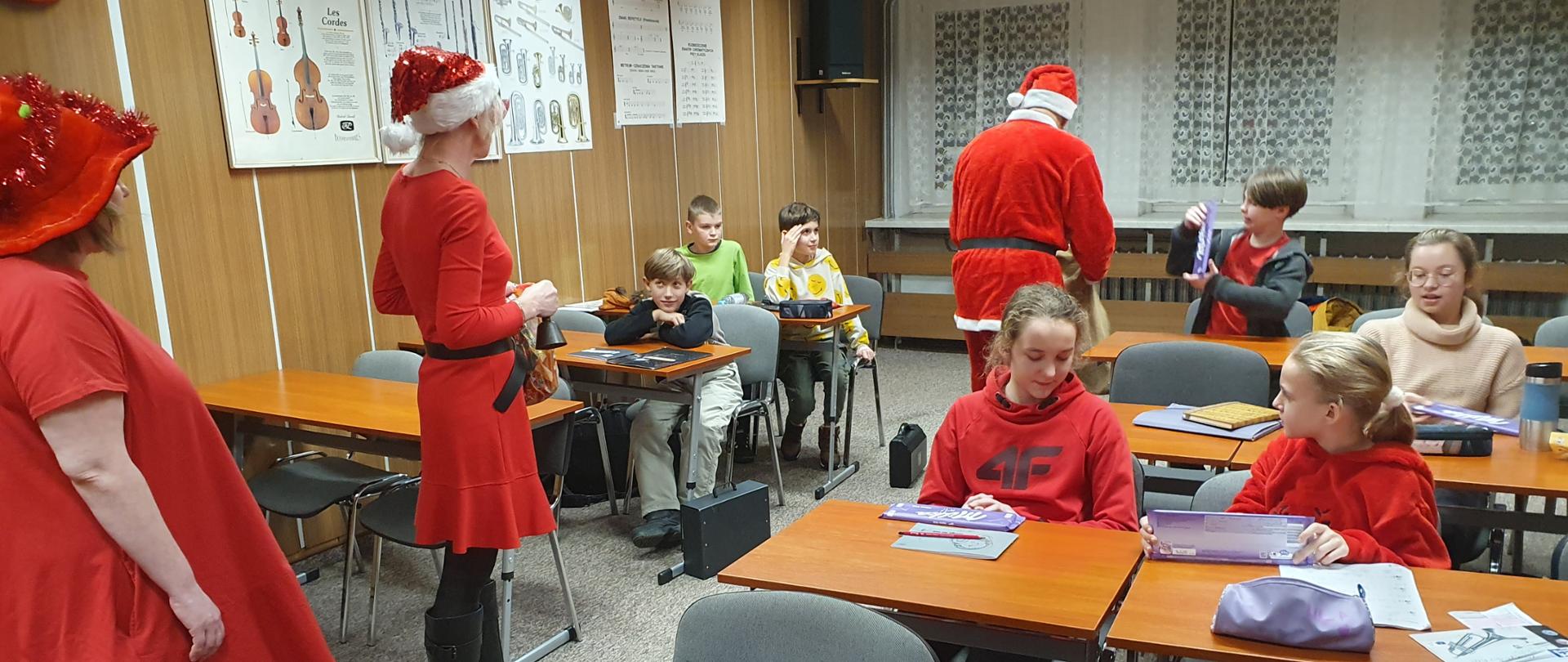 Mężczyzna w stroju Mikołaja i dwie kobiety w czerwonych strojach elfów rozdają dzieciom siedzącym w ławkach w sali lekcyjnej słodycze.
