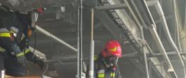 Zdjęcie przedstawia wnętrze hali produkcyjnej, w której doszło do zapalenia się ciągu wentylacyjnego. Widoczni strażacy w ubraniach specjalnych i z założonym sprzętem ochrony dróg oddechowych prowadzą działania przy demontażu urządzeń. 