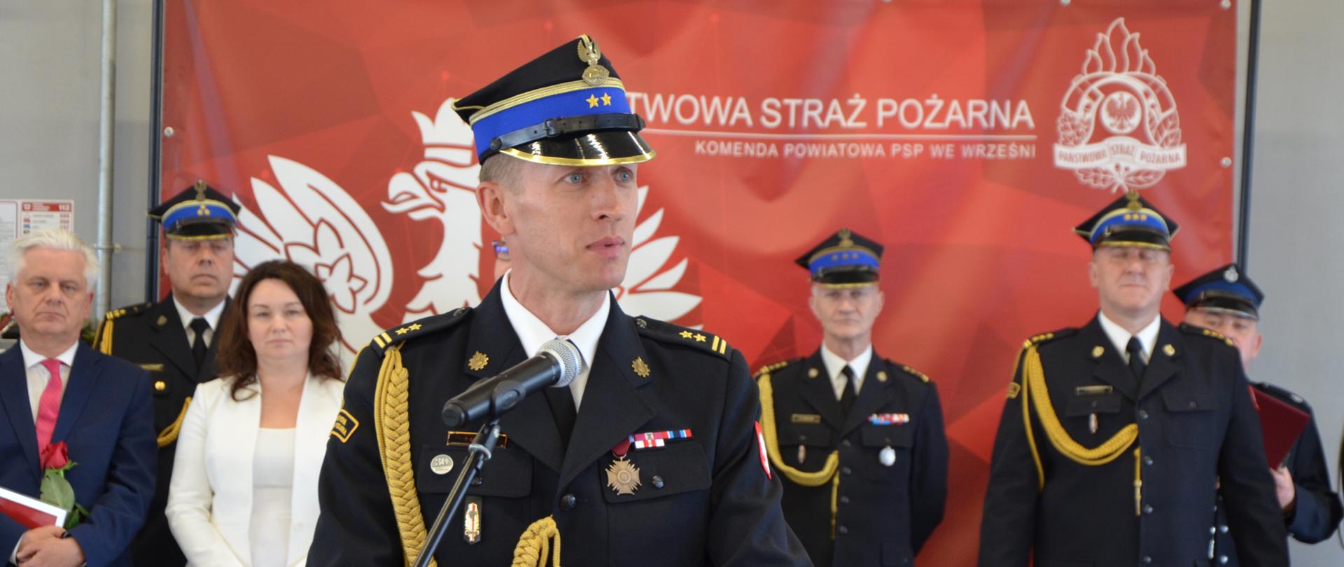strażak w rogatywce, w ciemnym mundurze ze złotym sznurem stoi przed mikrofonem, w tle grupa ludzi
