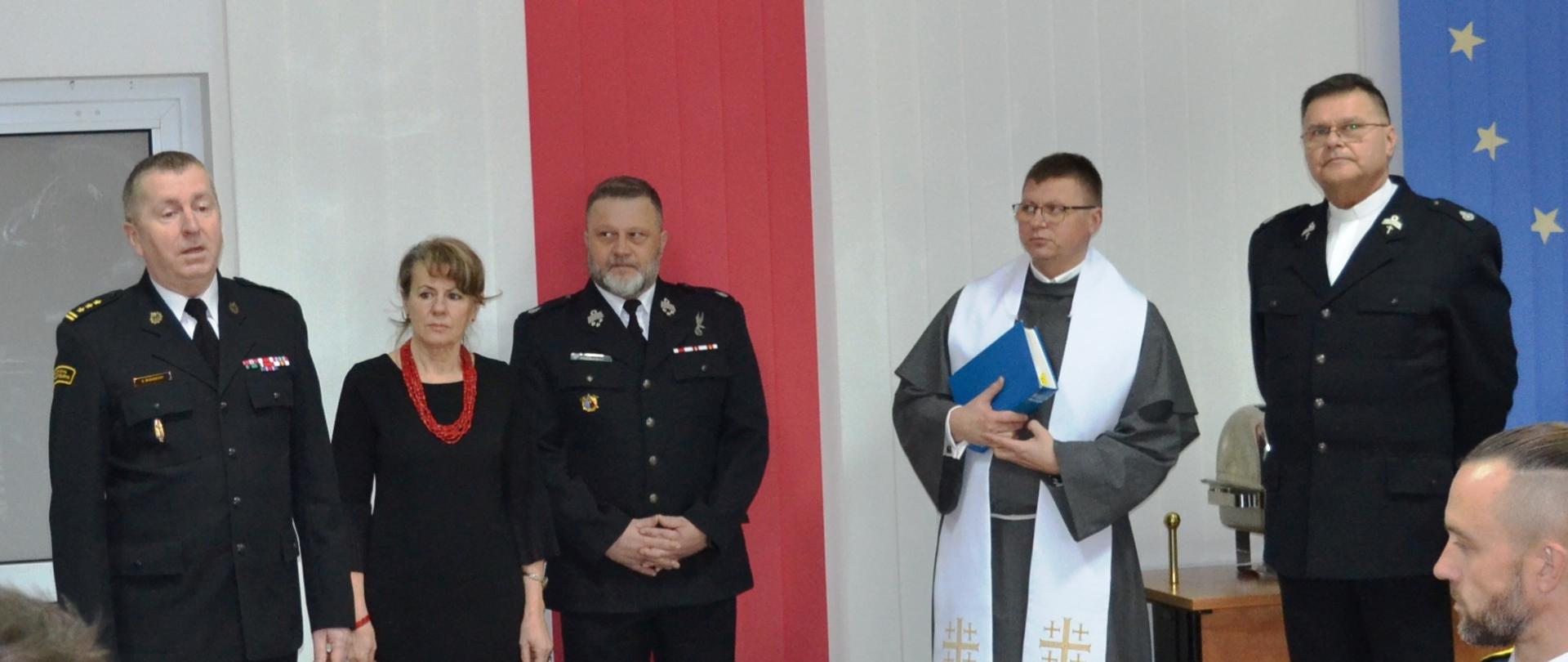 Komendant wita gości, obok niego stoją księża, kobieta cywil i dwójka strażaków.