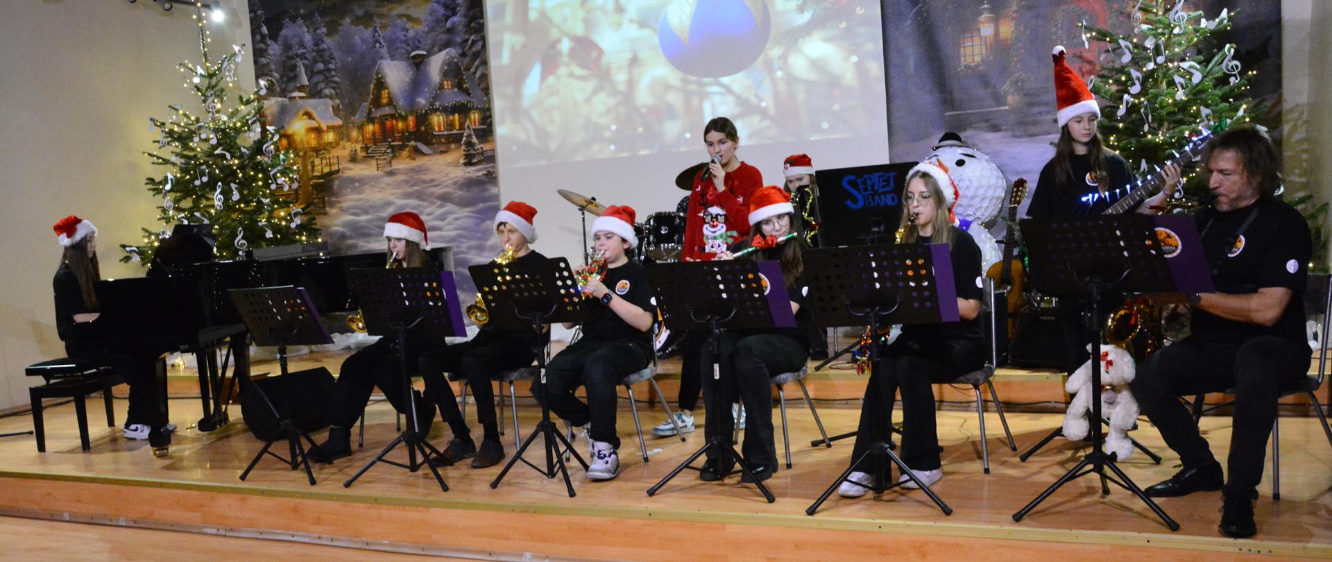 Uczniowie wraz z nauczycielem, od strony lewej dziewczynka przy fortepianie, dalej dziewczynka grająca na trąbce, dwóch chłopców grających na trąbce, dziewczynka grająca na flecie poprzecznym , dziewczynka grająca na saksofonie oraz mężczyzna grający na saksofonie, w głębi dziewczynka śpiewająca oraz grająca na gitarze w tle jeszcze jedna uczennica .