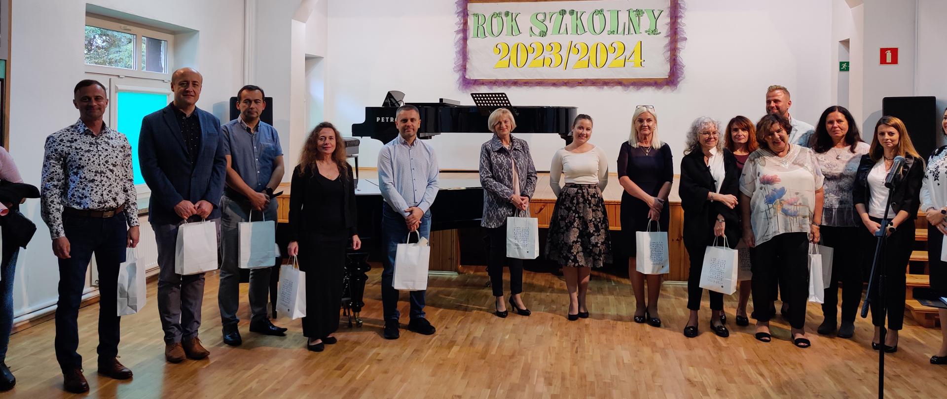 Zdjęcie przedstawiające nagrodzonych nauczycieli i pracowników szkoły przy scenie podczas koncertu w auli szkoły.