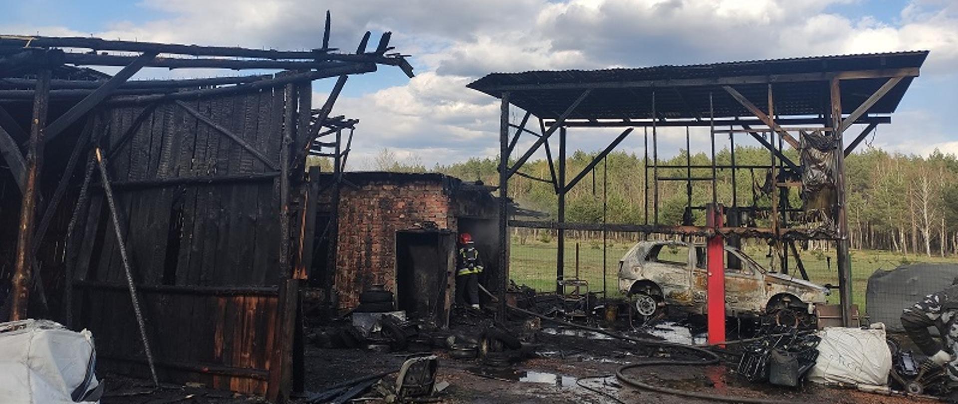 Spalone budynki gospodarcze. Na pierwszym planie z lewej strony nadpalona ściana drewniana budynku parterowego, bez dachu. Na drugim planie murowane pomieszczenie, w drzwiach którego stoi strażak prowadząc działania dogaszające pożar, Z prawej strony wiata pod którą stoi spalony samochód osobowy.