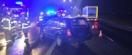 Zdjęcie przedstawia stojący na środku jezdni samochód osobowy po wypadku. Po jego lewej stronie znajdują się strażacy i wóz straży pożarnej. Za pojazdem karetka pogotowia, po prawej stronie TIR i pachołki odgradzające. 