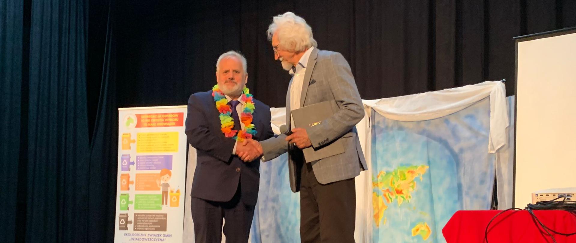 Dwóch mężczyzn w garniturach stoi na scenie i podaje sobie ręce 