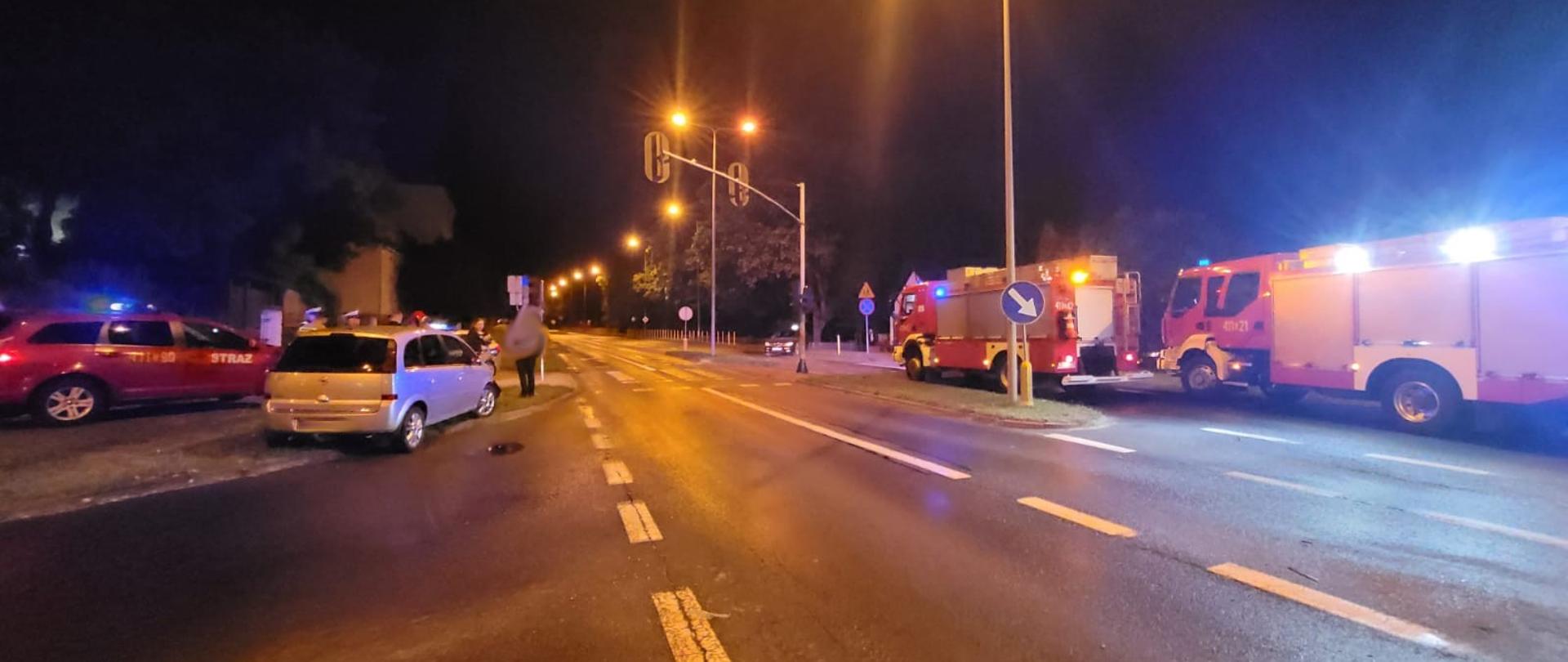 Pora nocna, zdjęcie przy sztucznym oświetleniu. Samochód osobowy po wypadku, stoi na krawędzi jezdni i pobocza. Wzdłuż jezdni widoczne dwa duże wozy strażackie. 