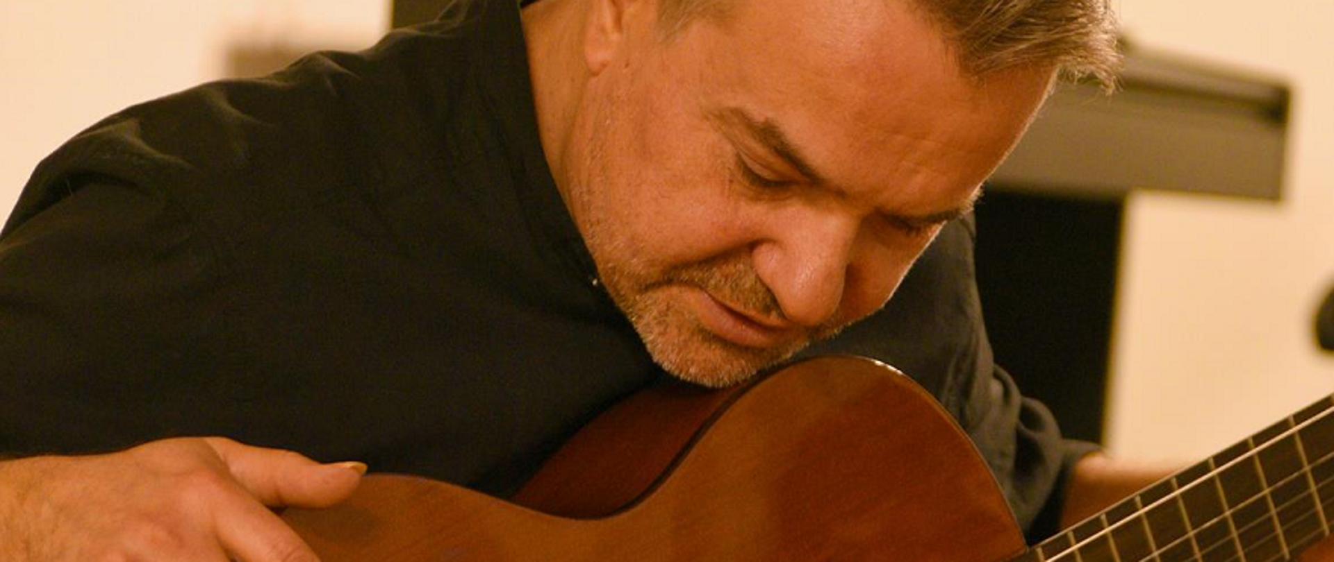 Zdjęcie przedstawia mężczyznę pochylonego nad trzymaną na kolanach gitarą na tle beżowej ściany.