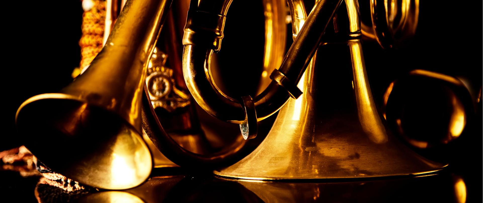 Zbliżenie na błyszczące mosiężne instrumenty dęte blaszane, w tym róg i trąbkę, ustawione na ciemnym tle; światło podkreśla złocisty kolor metalu i detale konstrukcyjne.