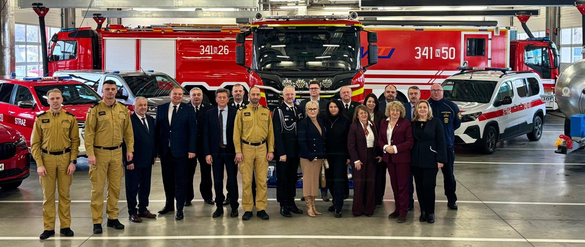 Zdjęcie przedstawia wnętrze dużej remizy strażackiej. W przestronnym, dobrze oświetlonym garażu stoją dwa masywne, czerwone wozy strażackie. Mają charakterystyczne białe i żółte oznaczenia oraz numery taktyczne umieszczone z boku. Na jednym z nich widnieje napis „STRAŻ”, co wskazuje, że to polskie pojazdy pożarnicze.
Przed samochodami stoi grupa kilkunastu osób ustawionych w dwóch rzędach. Część z nich to strażacy w ciemnych, służbowych mundurach. Pozostali są ubrani bardziej formalnie — w garnitury, płaszcze lub eleganckie ubrania — co sugeruje, że mogą to być goście, przedstawiciele władz lub uczestnicy oficjalnej wizyty.
Wszyscy pozują do wspólnego zdjęcia, stojąc blisko siebie i patrząc w stronę aparatu. Atmosfera wygląda na uroczystą, ale spokojną. W tle, oprócz pojazdów, widać elementy konstrukcji budynku: metalowe belki, lampy sufitowe i instalacje wentylacyjne.