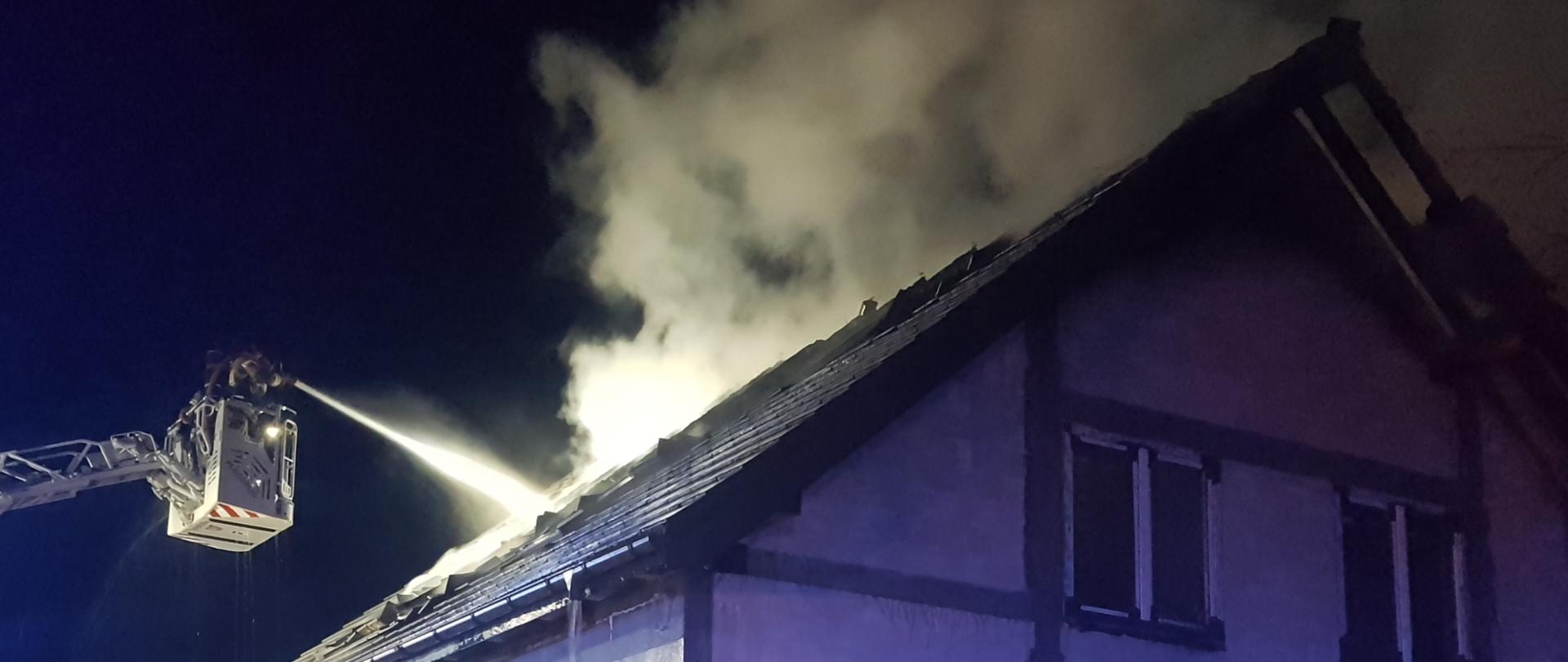 Nowo wybudowany dom mieszkalny. Z dachu wydobywa się dym. Nad budynkiem widoczny jest kosz podnośnika z którego strażacy gaszą pożar na dachu.