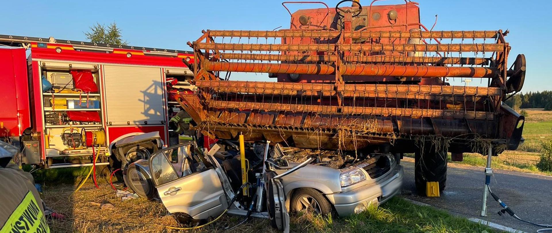Zdjęcie uwidacznia kombajn zbożowy pod jego zespołem tnącym zgnieciony samochód osobowy w tle samochód pożarniczy 
