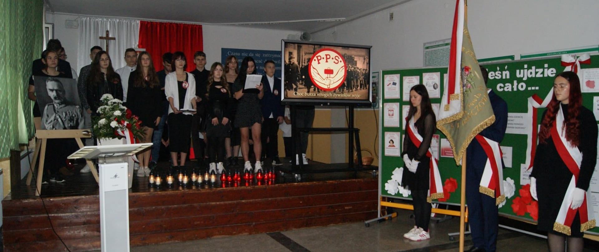 Szerokie ujęcie uroczystości. Na scenie grupa uczniów recytuje tekst, obok nich poczet sztandarowy. Na ekranie widoczne historyczne logo PPS (Polska Partia Socjalistyczna) na tle archiwalnej fotografii.