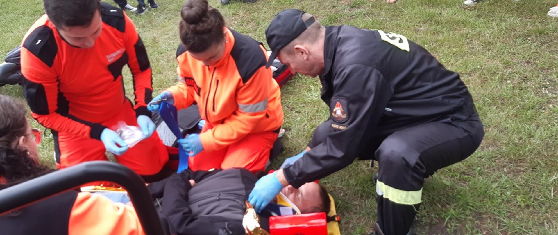 Na zdjęciu strażak oraz ratownicy medyczni udzielający pomocy przedmedycznej osobie poszkodowanej w ramach ćwiczeń.W tle obserwatorzy.