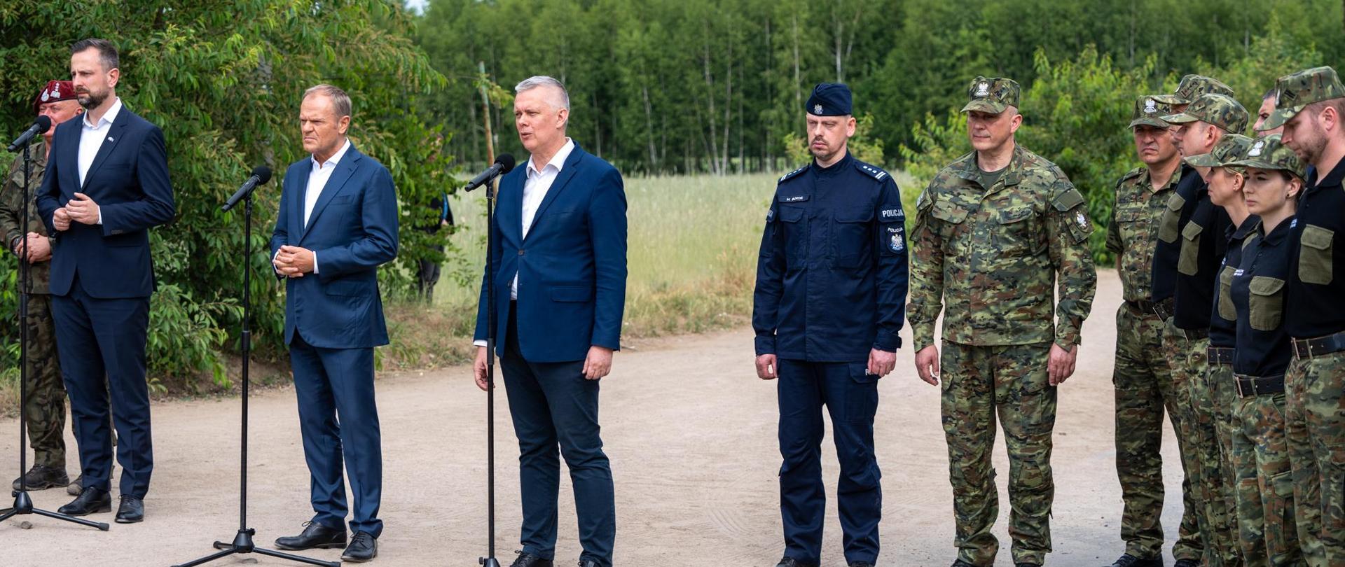 Od lewej wicepremier Kosiniak-Kamysz, premier Tusk i minister Siemoniak stoją przy mikrofonach. Po prawej stronie ministra Siemoniaka grupa funkcjonariuszy straży granicznej. W tle łąka i las. 