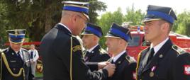 Zdjęcie przedstawia Zastępcę Kujawsko-Pomorskiego Komendanta Wojewódzkiego Państwowej Straży Pożarnej starszego brygadiera Artura Przybylskiego przypinającego medal funkcjonariuszom Komendy. Po lewo do gratulacji przygotowuje się Komendant Powiatowy. Wszyscy ubrani w stroje galowy. Słonce odbija się w otokach czapek rogatywek. W te zabytkowy pojazd gaśniczy.