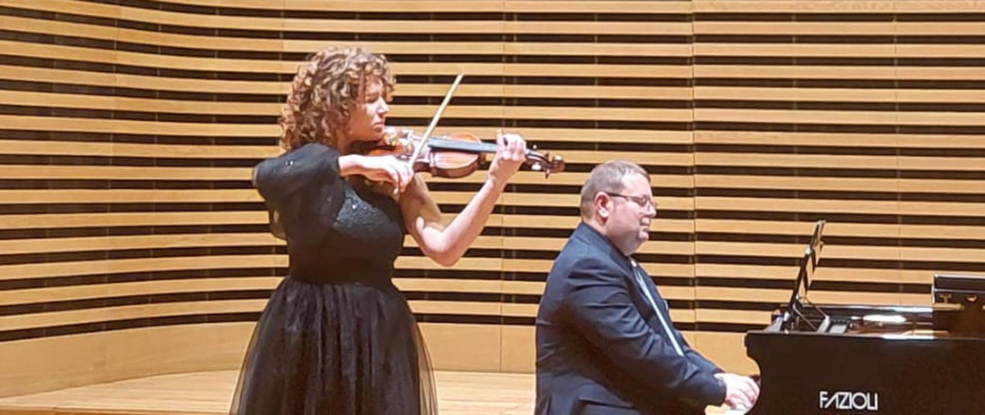 Dziewczyna gra na skrzypcach, mężczyzna akompaniuje na fortepianie.