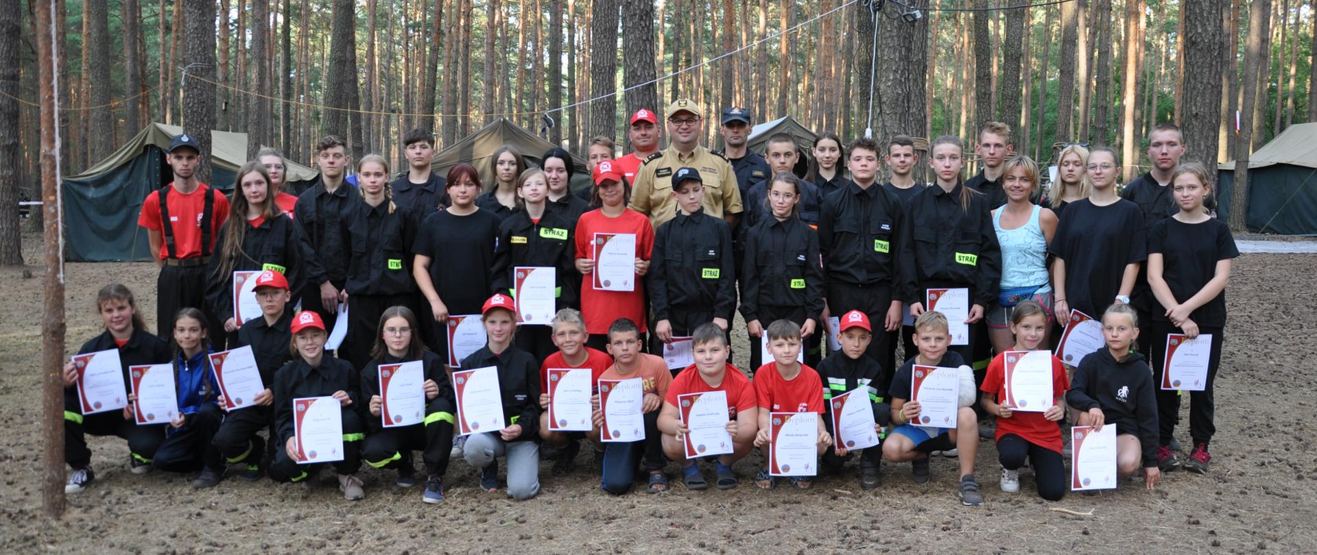 grupa dzieci i młodzieży stoi , pierwszy rząd klęczy, każdy z nich trzyma w ręku dyplom, w środku grupy stoi mężczyzna w jasnym mundurze i czapeczce, strażak, zdjecie zrobione w lesie