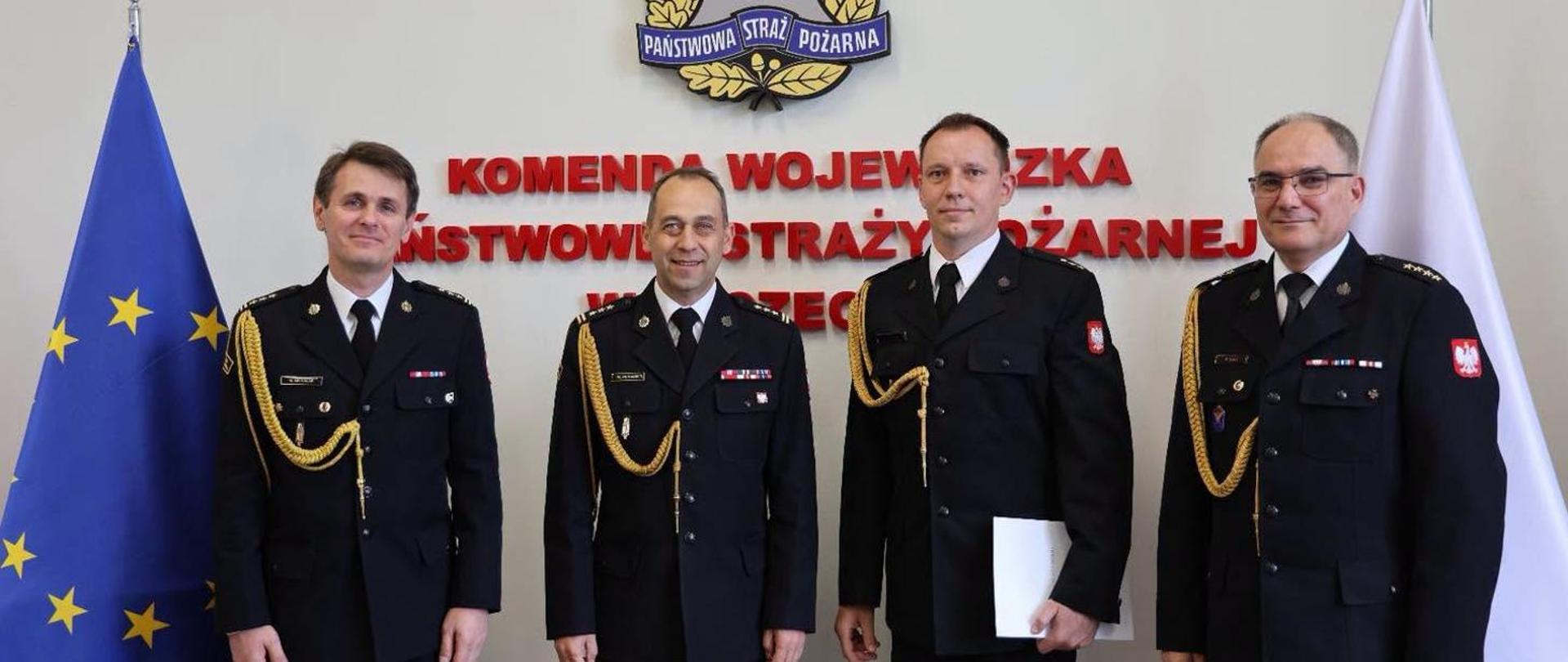 Na zdjęciu widać czterech strażaków ubranych w mundury wyjściowe, koloru czarnego ze sznurami galowymi koloru żółtego. Strażacy stoją na tle napisu Komeda Wojewódzka Państwowej Straży pożarnej w Szczecinie. z lewej strony widać flagę unijną koloru niebieskiego, z prawej flagę polski koloru biało-czerwonego