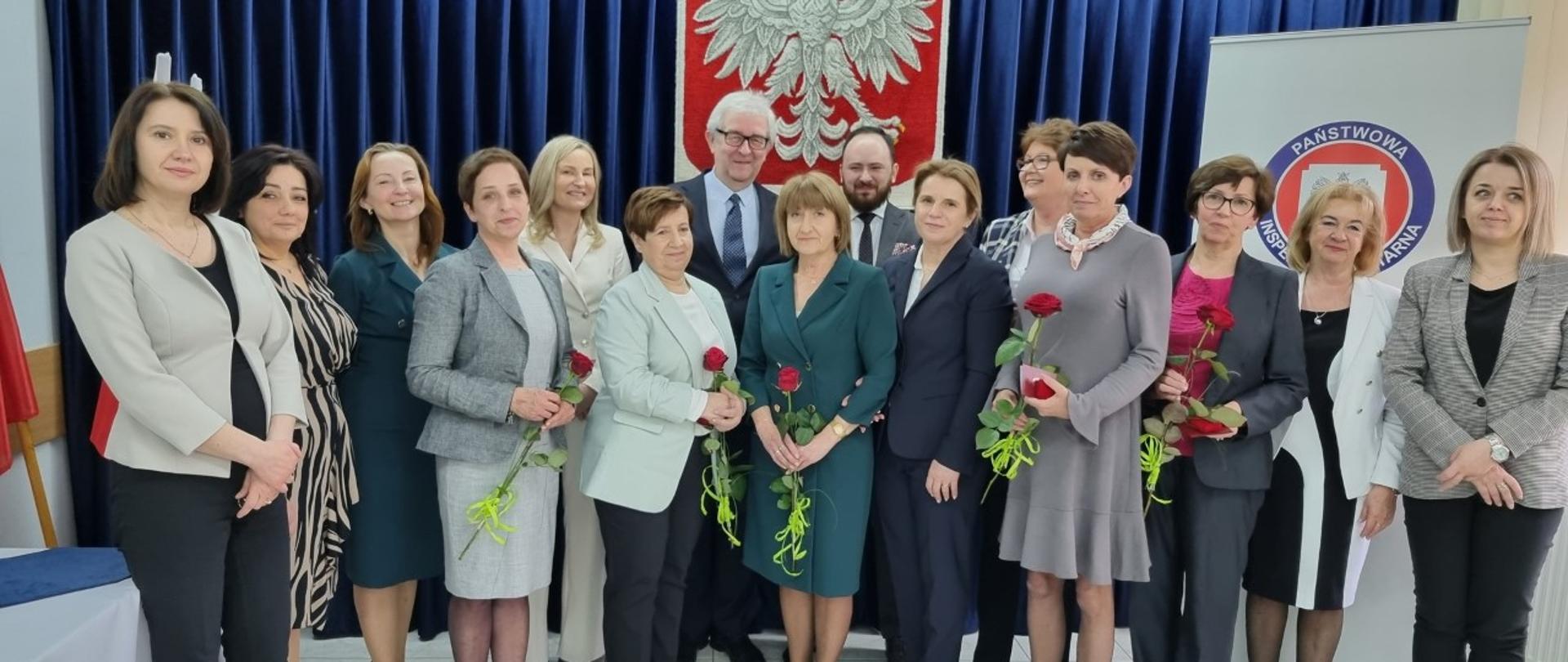 Od lewej: kobiet w jasnej marynarce i ciemnych spodniach, kobieta w sukience w czarno-beżowe pasy, kobieta w zielonej sukience, przed nią kobieta w jasnej sukience i szarej marynarce z różą w dłoni, za nią kobieta w białym garniturze, przed nią kobieta w jasnoszarej marynarce i ciemnych spodniach z różą w dłoni, za nią mężczyzna w ciemnym garniturze, w okularach, przed nim kobieta w zielonej sukience z różą w dłoni, za nią brodaty mężczyzna, przed nim kobieta w granatowym garniturze, obok kobieta w szarej sukience z różą w dłoni, za nią kobieta w okularach w marynarce w czarno-białą kratę, obok kobieta w różowej bluzce i ciemno-szarym garniturze, obok kobieta w biało-czarnej sukience, obok kobieta w szarej marynarcie i czarnych spodniach. Z tyłu na granatowej kurtynie godło Polski