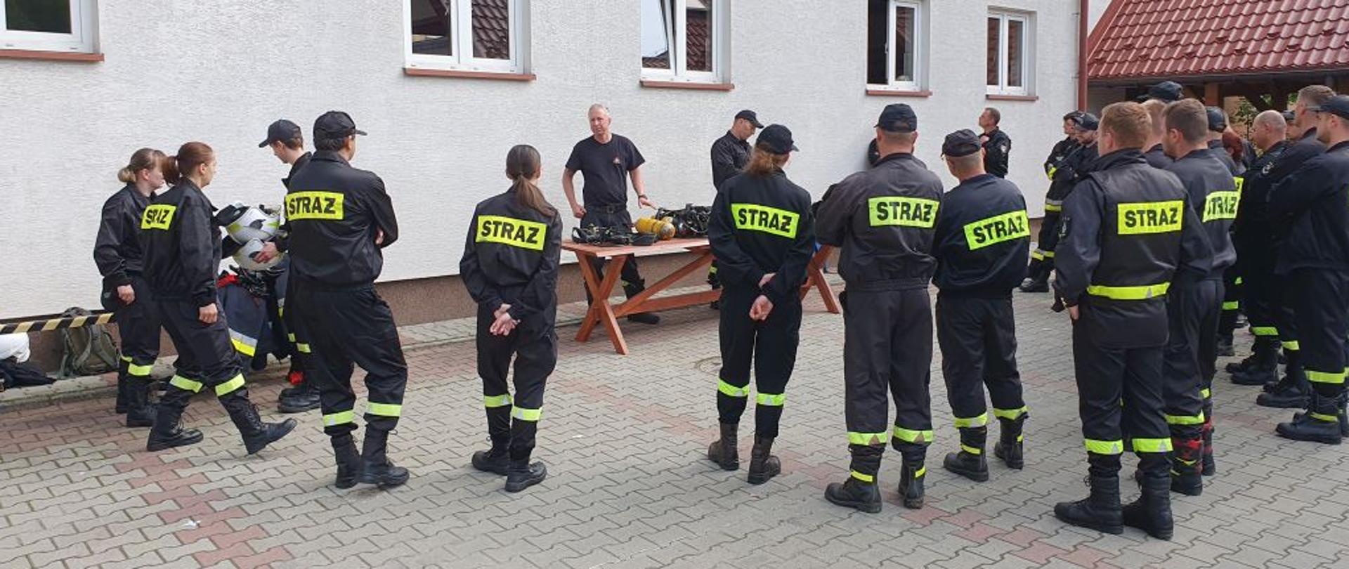 Na zdjęciu widać dwóch strażaków z PSP którzy stoją przy stole na którym leżą butle stalowe oraz noszaki i maski. Na około nich stoją strażacy z OSP. Wszyscy ubrani są w ubrania koszarowe. Zdjęcie wykonane jest na tylnim placu komendy. 